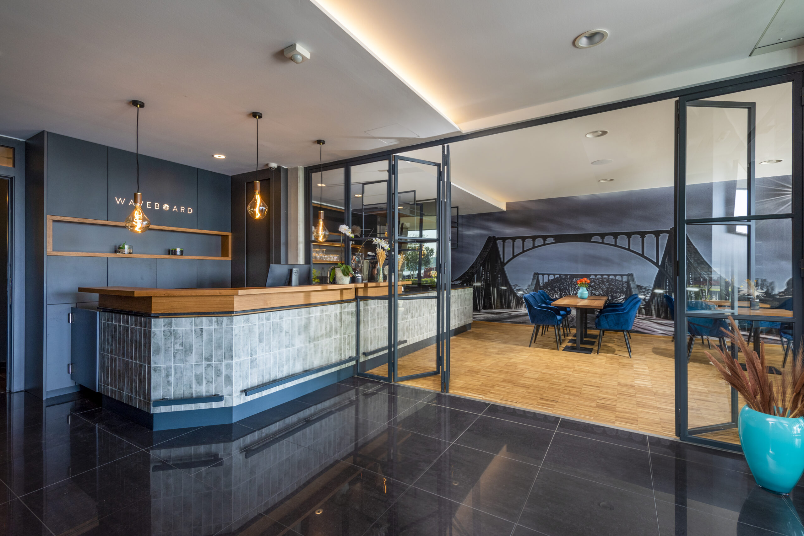 Empfangsbereich des WAVE Hotel Potsdam mit moderner Rezeption aus Holz und Fliesen, stilvollen Hängelampen, Glasabtrennung und Blick in einen hellen Konferenzraum mit Wandbild der Glienicker Brücke.