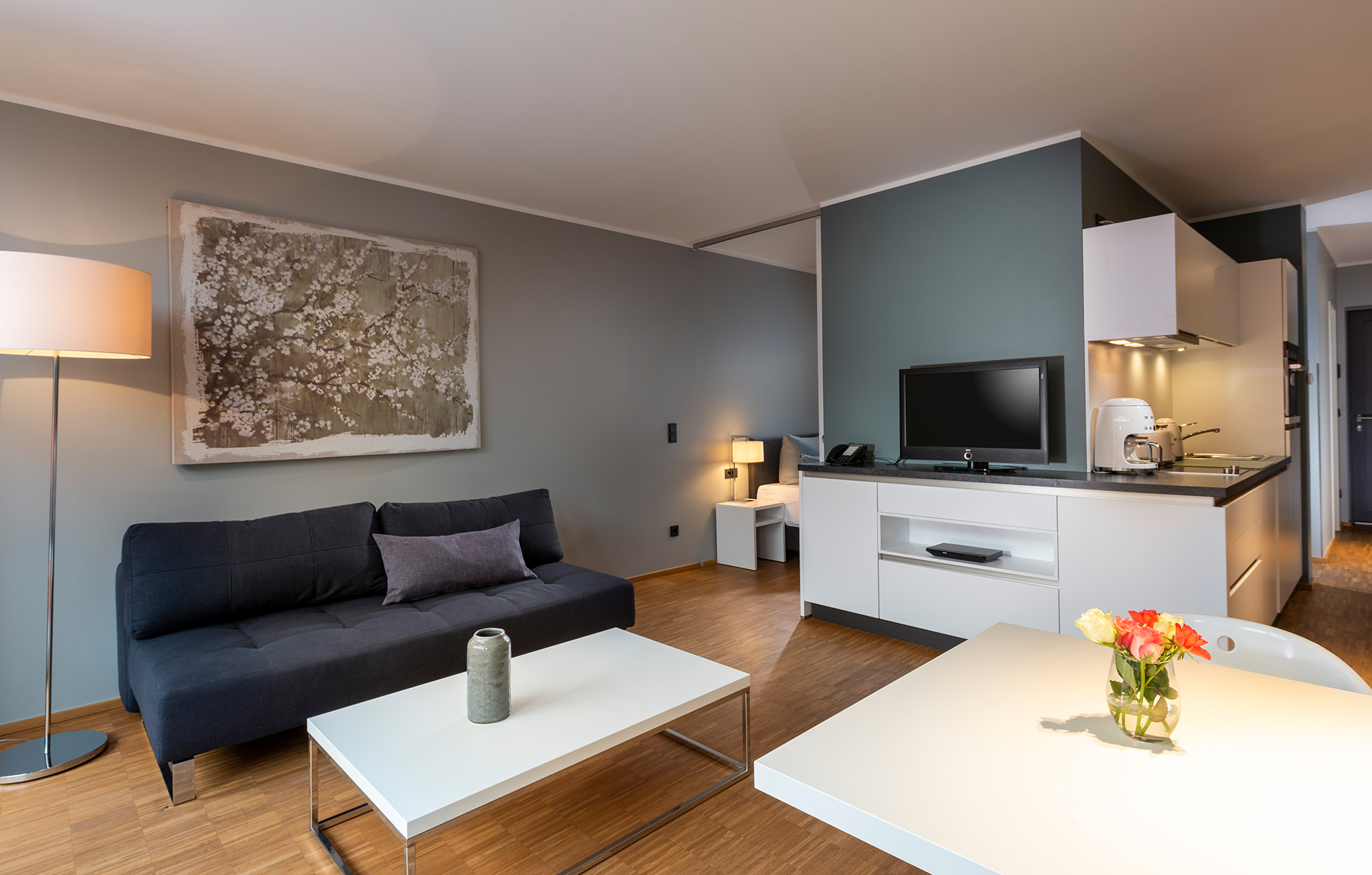 Modernes Apartment im WAVE Hotel Potsdam mit Wohnbereich, Sofa und Couchtisch, offener Küchenzeile mit TV, Esstisch mit Blumen und Blick ins angrenzende Schlafzimmer.