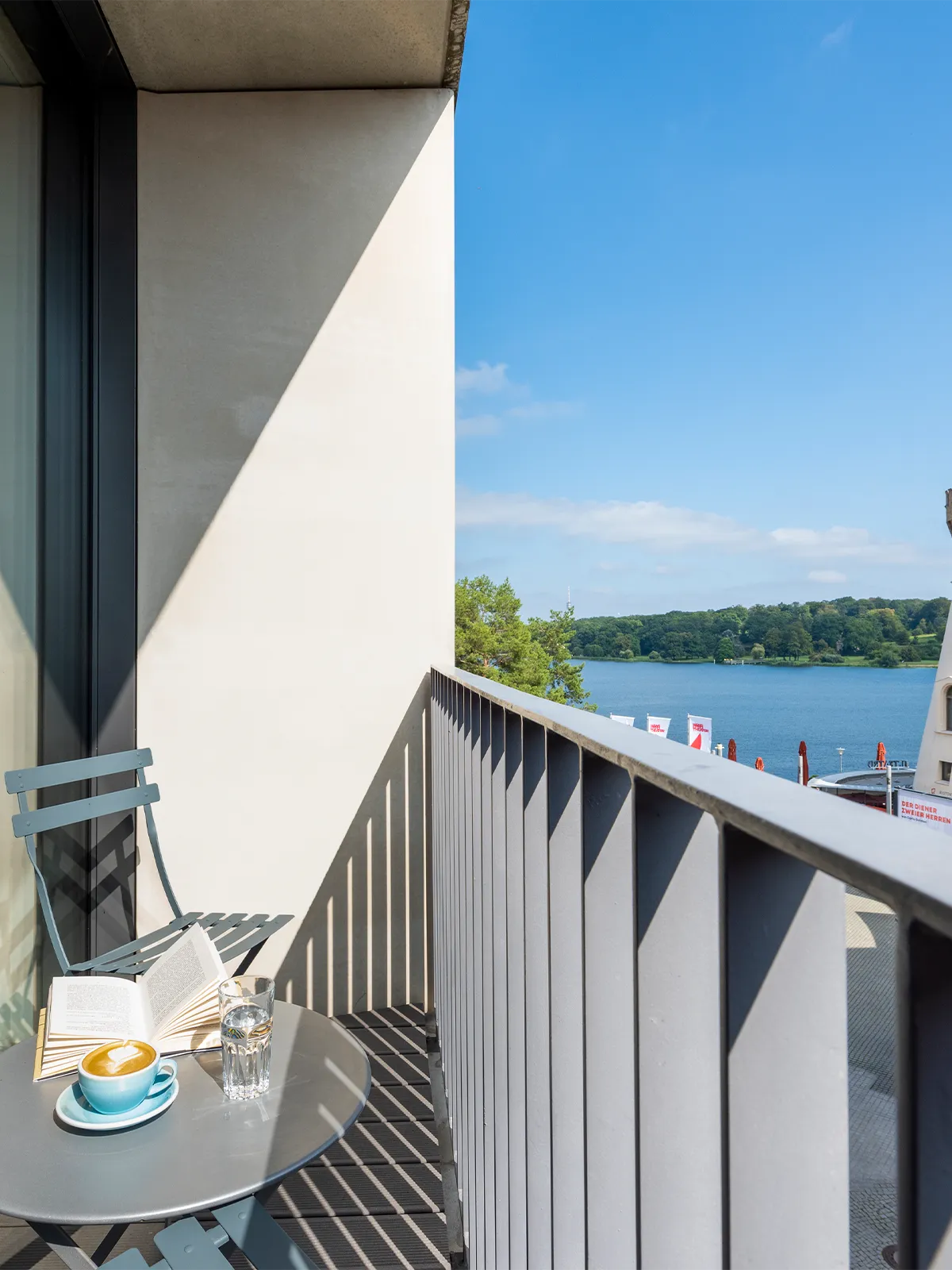 Kleiner Balkon im WAVE Hotel Potsdam mit Sitzgelegenheit, Kaffee und Buch auf einem runden Tisch, sowie Blick auf die Havel und grüne Uferlandschaft bei blauem Himmel.