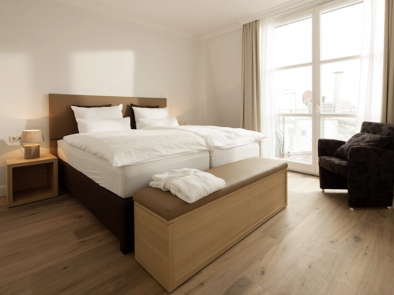 Blick in das Apartment Maison Penthouse N°5 der Villa Vie auf Norderney. Zu sehen ist ein bequemes Doppelbett mit hellem lichtdurchflutetem Ambiente und einem Sessel.