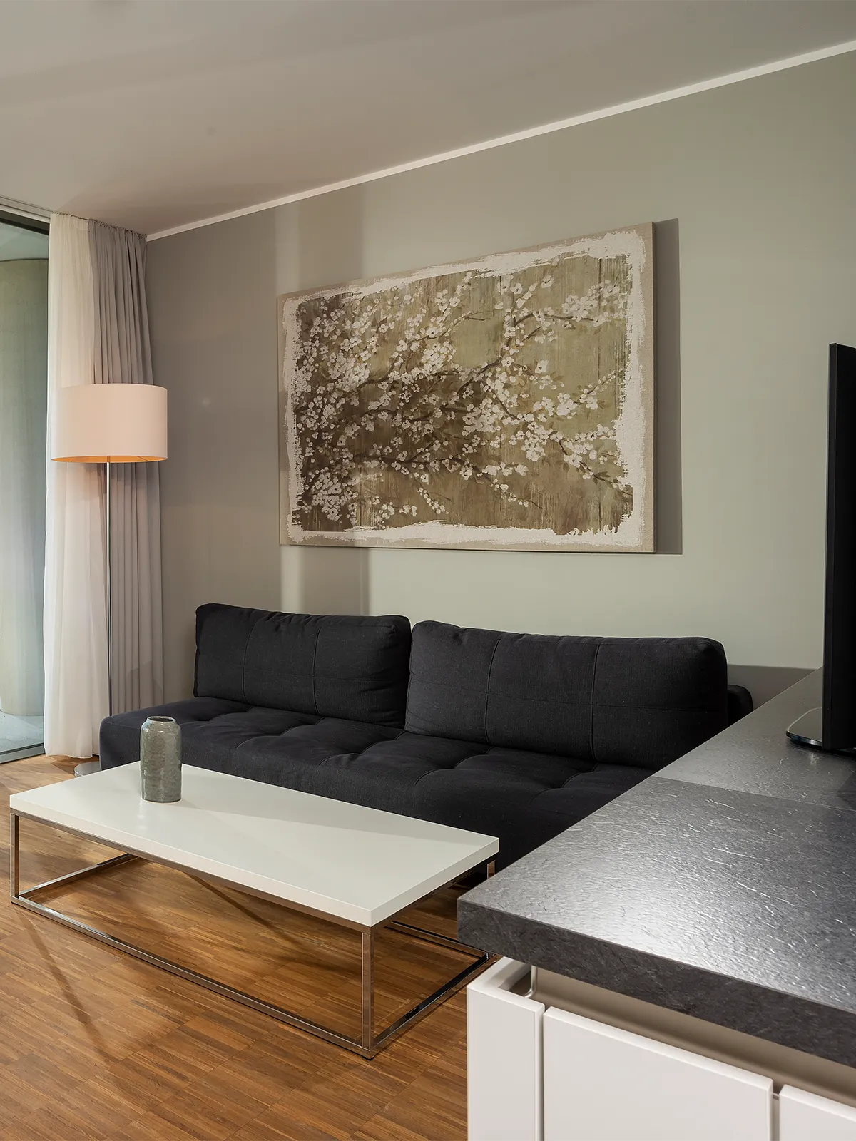 Moderner Wohnbereich im WAVE Hotel Potsdam mit dunklem Sofa, weißem Couchtisch, dekorativer Stehlampe und großformatigem Wandbild auf heller Wand.