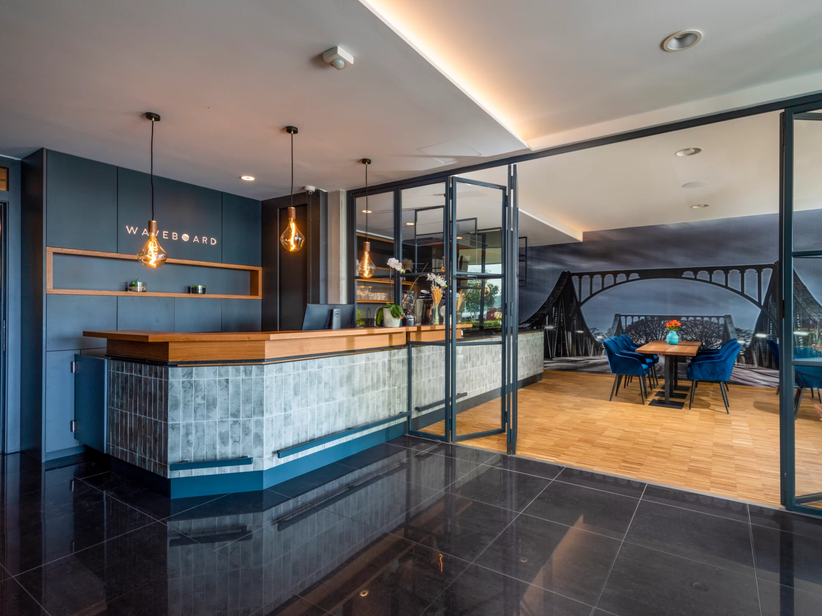 Moderner Empfangsbereich eines Hotels mit stilvollem Tresen aus Holz und Fliesen, darüber drei dekorative Pendelleuchten; rechts durch Glaswände abgetrennter Lounge- oder Frühstücksbereich mit Holzfußboden, blauen Sesseln und großem Wandbild einer Brücke.