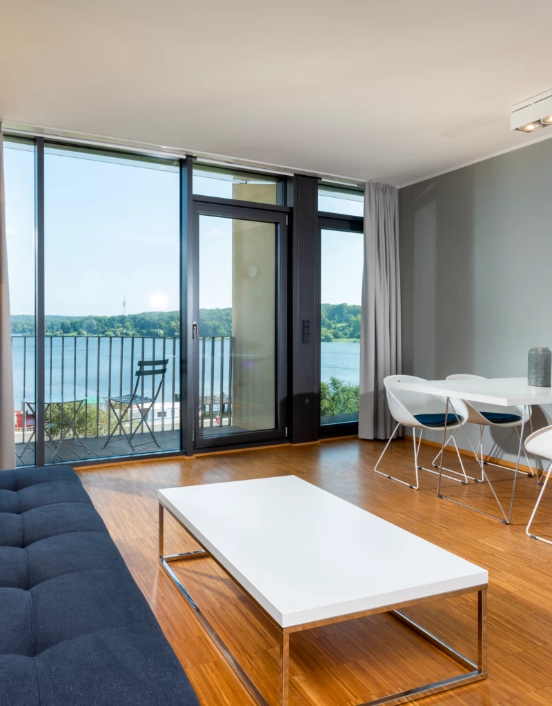 Heller Wohn- und Essbereich im WAVE Hotel Potsdam mit modernem Sofa, weißem Couchtisch und Esstisch sowie bodentiefen Fenstern mit Ausblick auf die Havel und grüne Uferlandschaft.